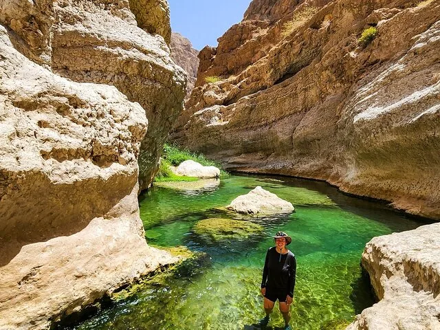 A smiling person wading in the crystal-clear green water of Wadi Shab canyon.