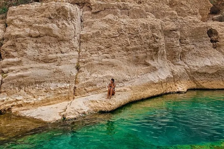Una persona sentada en una repisa rocosa sobre una poza verde cristalina en Wadi Shab.